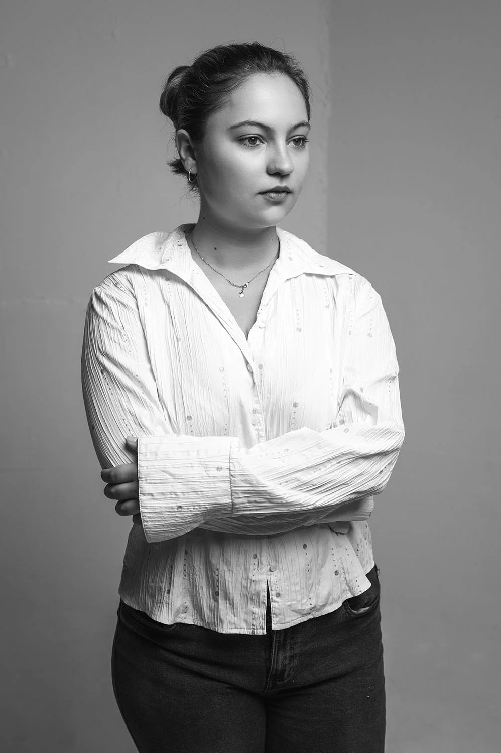 Frau, die eine weiße Bluse trägt, steht vor einer weißen Wand. Sie hat die Arme vor sich verschränkt und schaut verträumt an der Kamera vorbei.