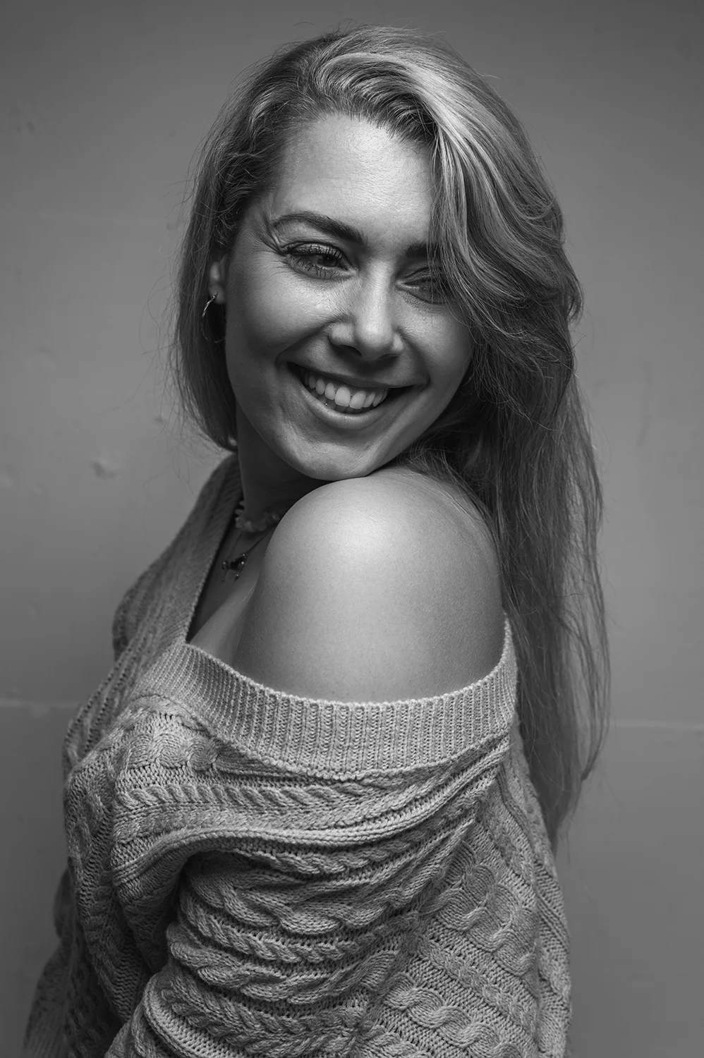 Blonde Frau trägt einen gestrickten Pullover und schaut grinsend über die Schulter zur Seite.
