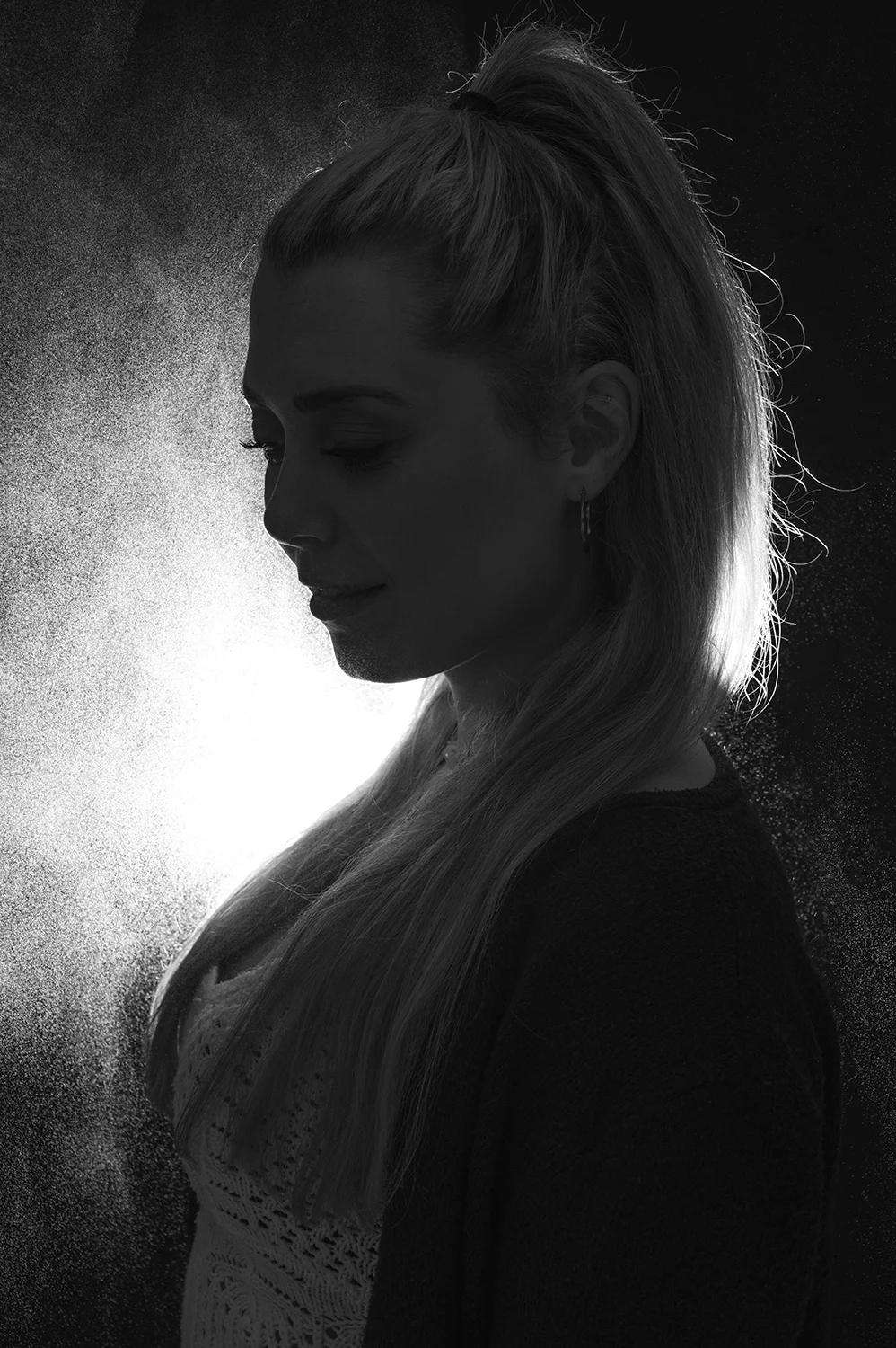 Blonde Frau steht vor einem dunklen Hintergrund während ein helles Licht und ein Sprühnebel zu sehen sind
